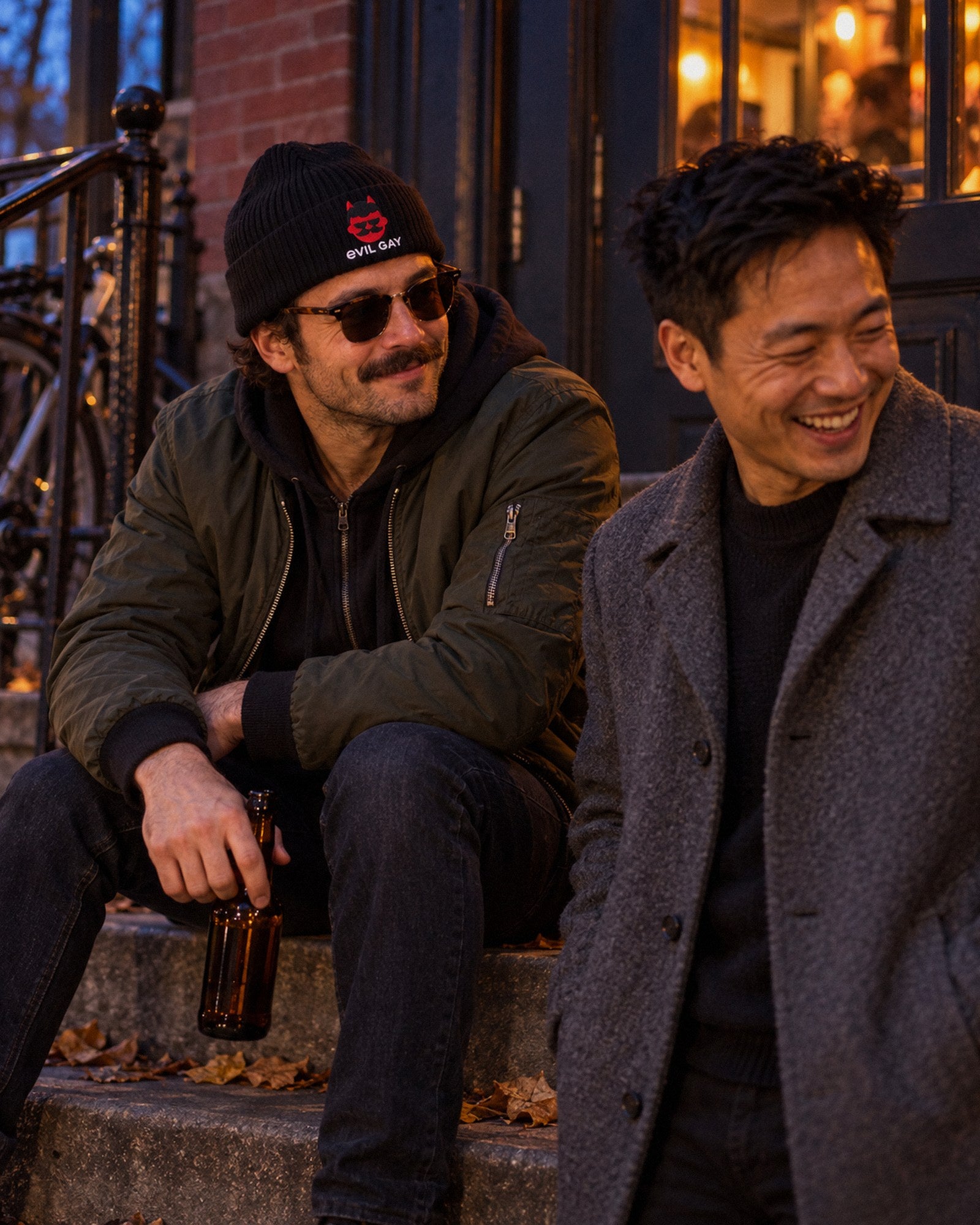 A mate in the black Evil Gay Beanie sitting on a bar stoop at dusk while his friend laughs
