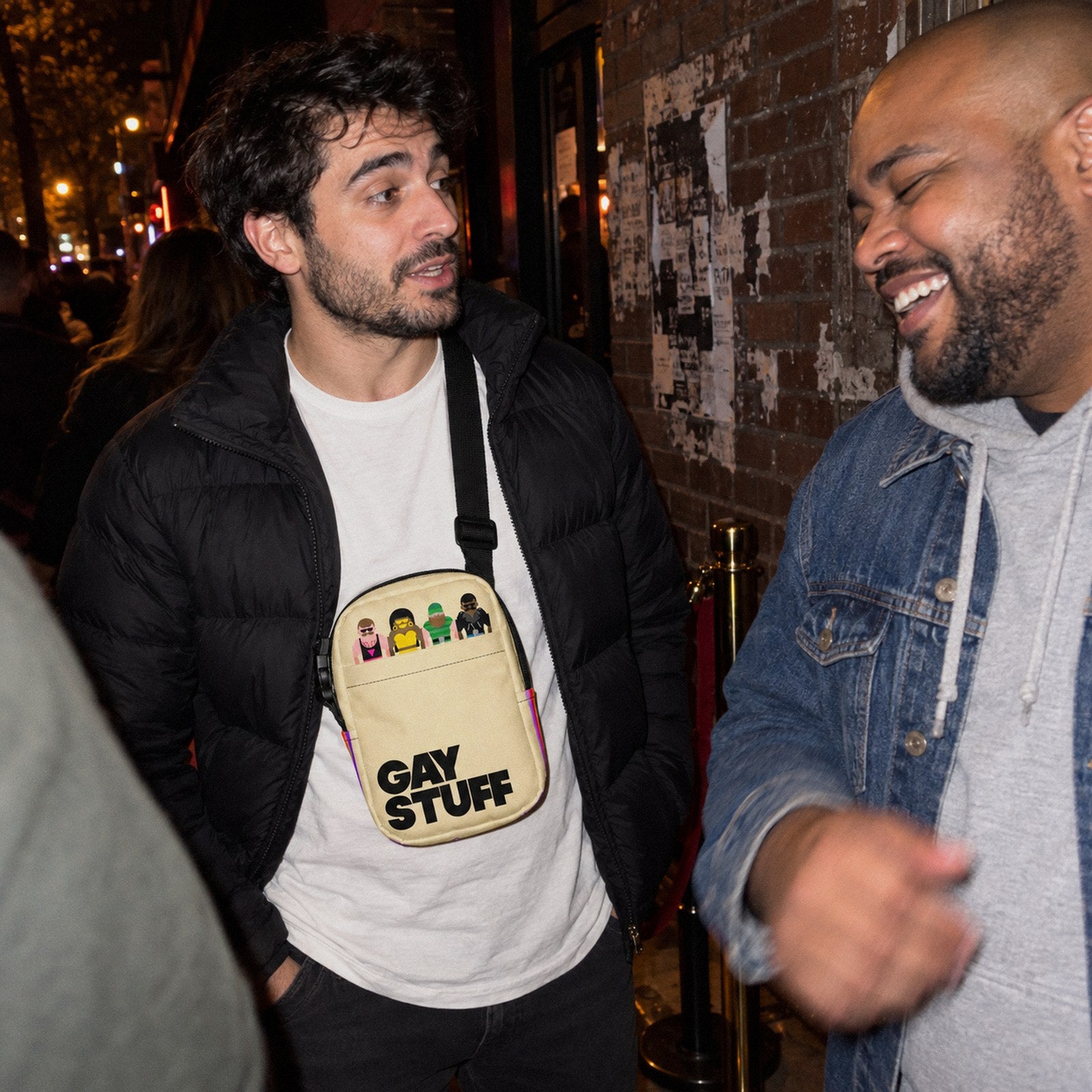 Two friends talking in line outside a bar at night, one wearing the Gay Stuff Crossbody Bag across his chest
